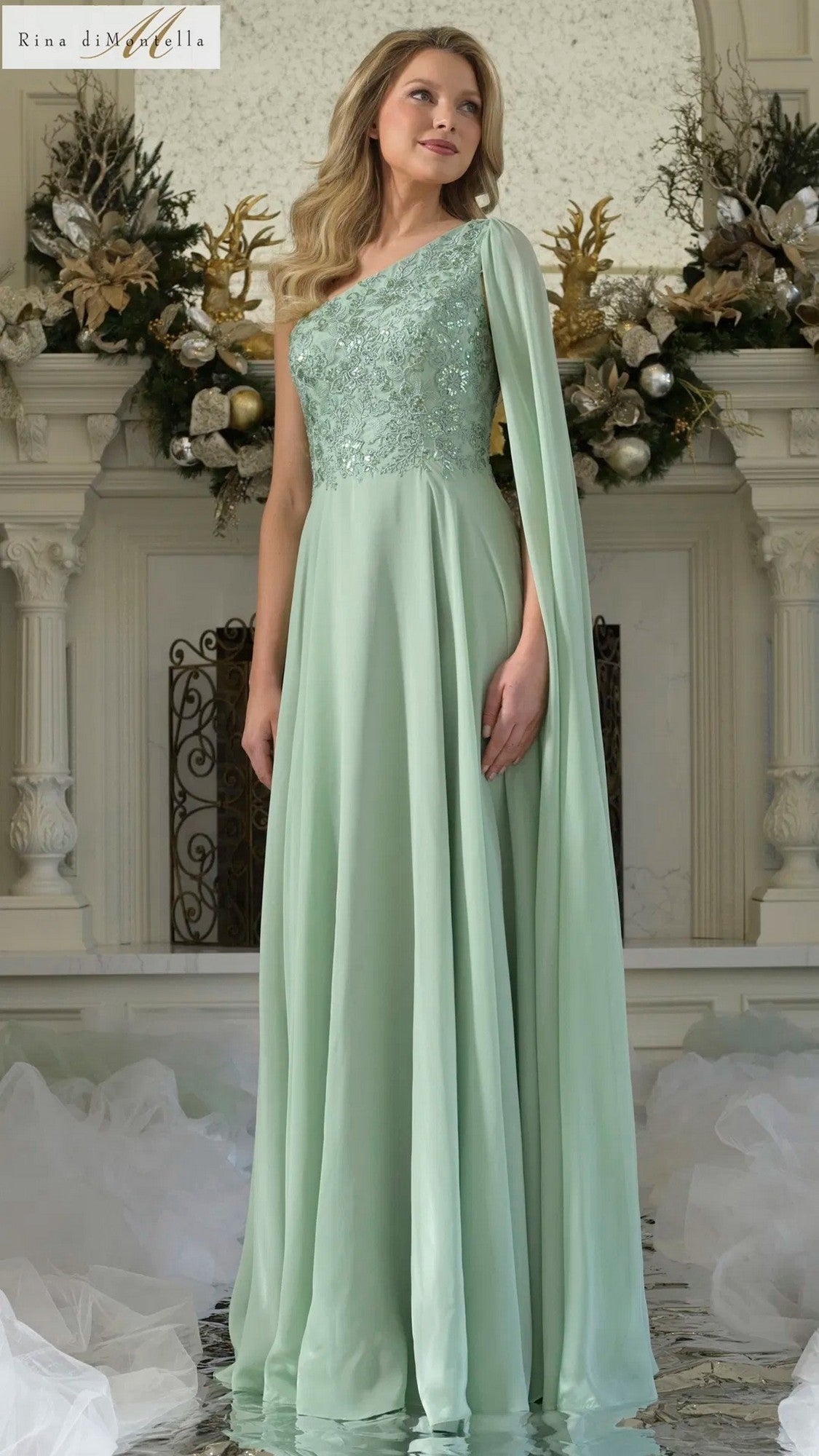 A woman stands indoors by an ornate gold and white fireplace, wearing the Rina Di Montella RD3354 long formal dress in light sage, featuring a one-shoulder design and a flowing cape.